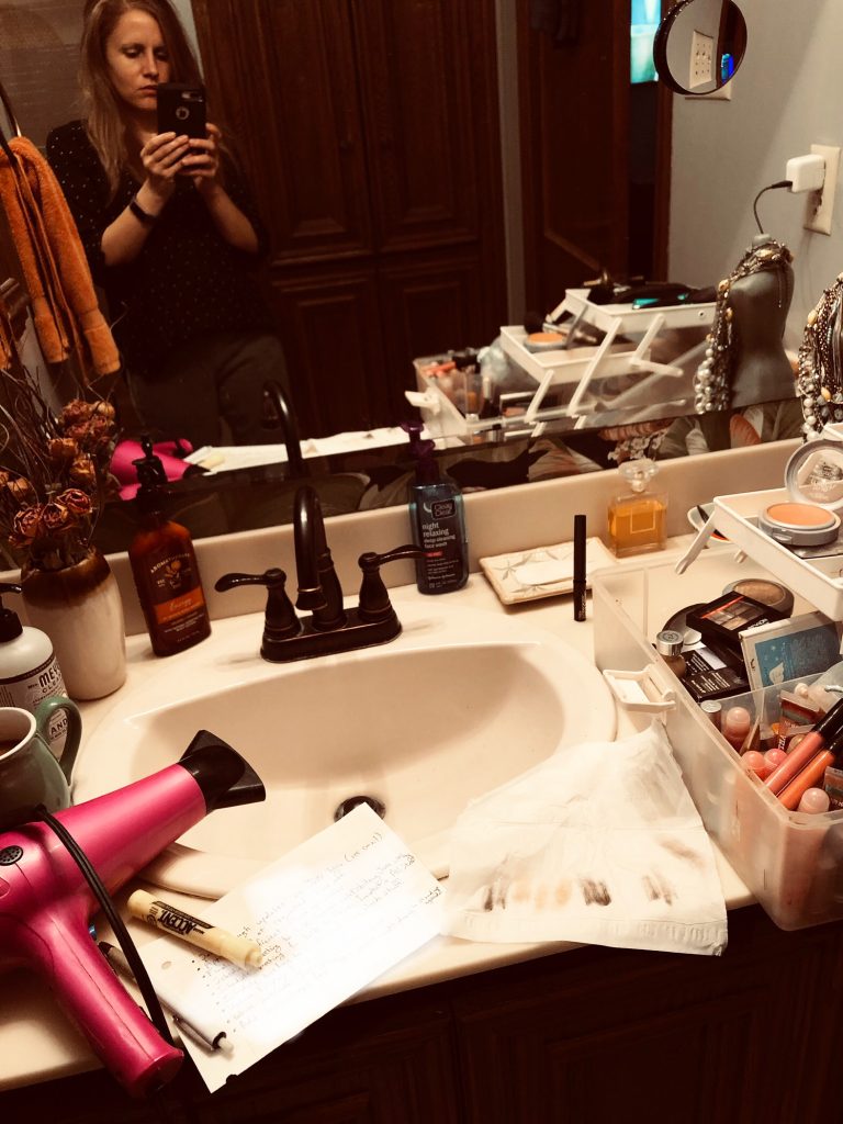 The crowded vanity of a woman mutli-tasker getting ready for the day. She pauses to reflect and is seen in the mirror taking a picture of it with her phone.