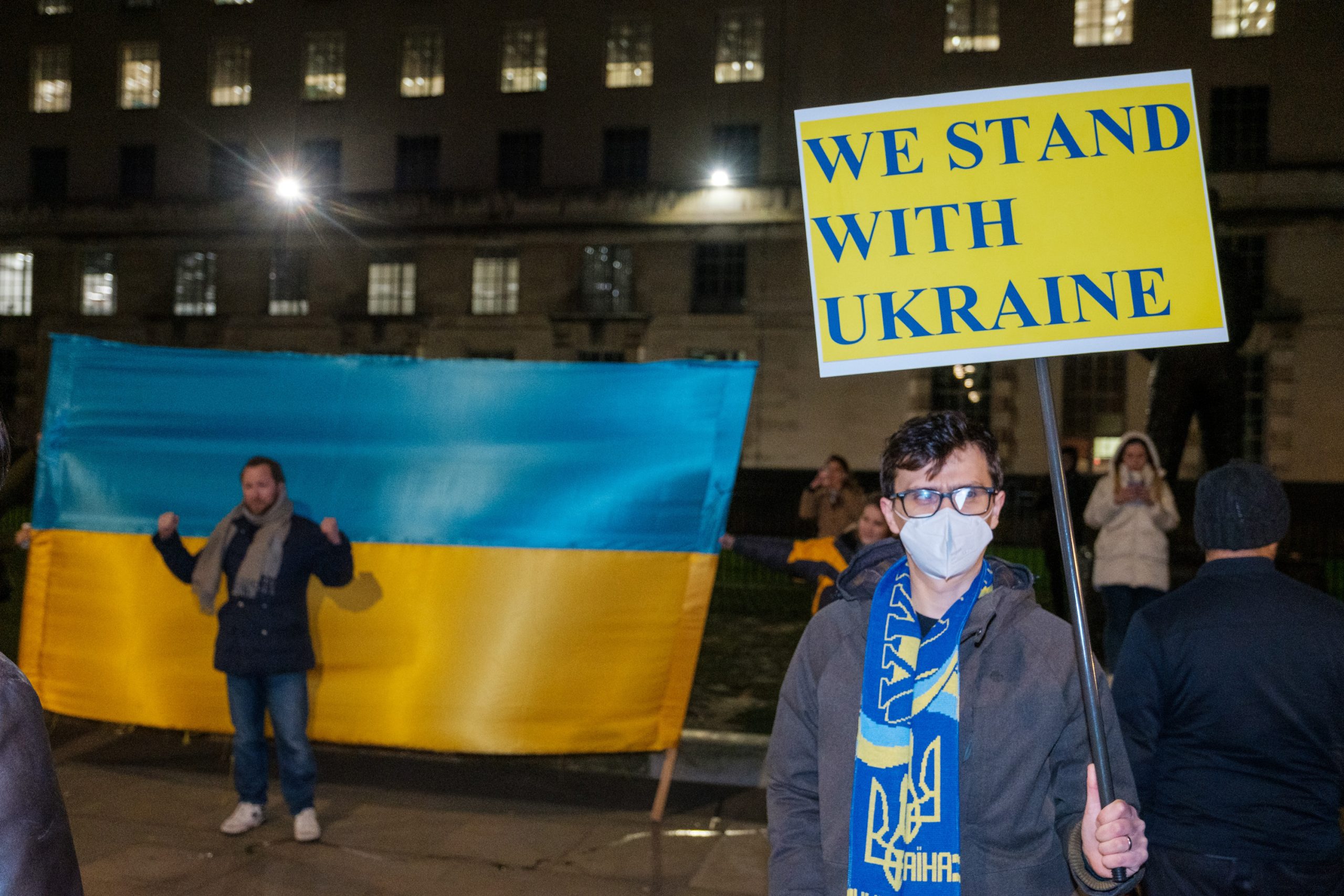 Men protesting in the UK with a large Ukrainian flag and a sigh that says WE STAND WITH UKRAINE. Image by Ehimetalor Akhere Unuabona.